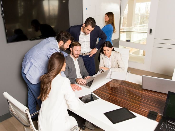 Six coworkers in a modern office huddle around a laptop on a white conference table, collaborating closely.