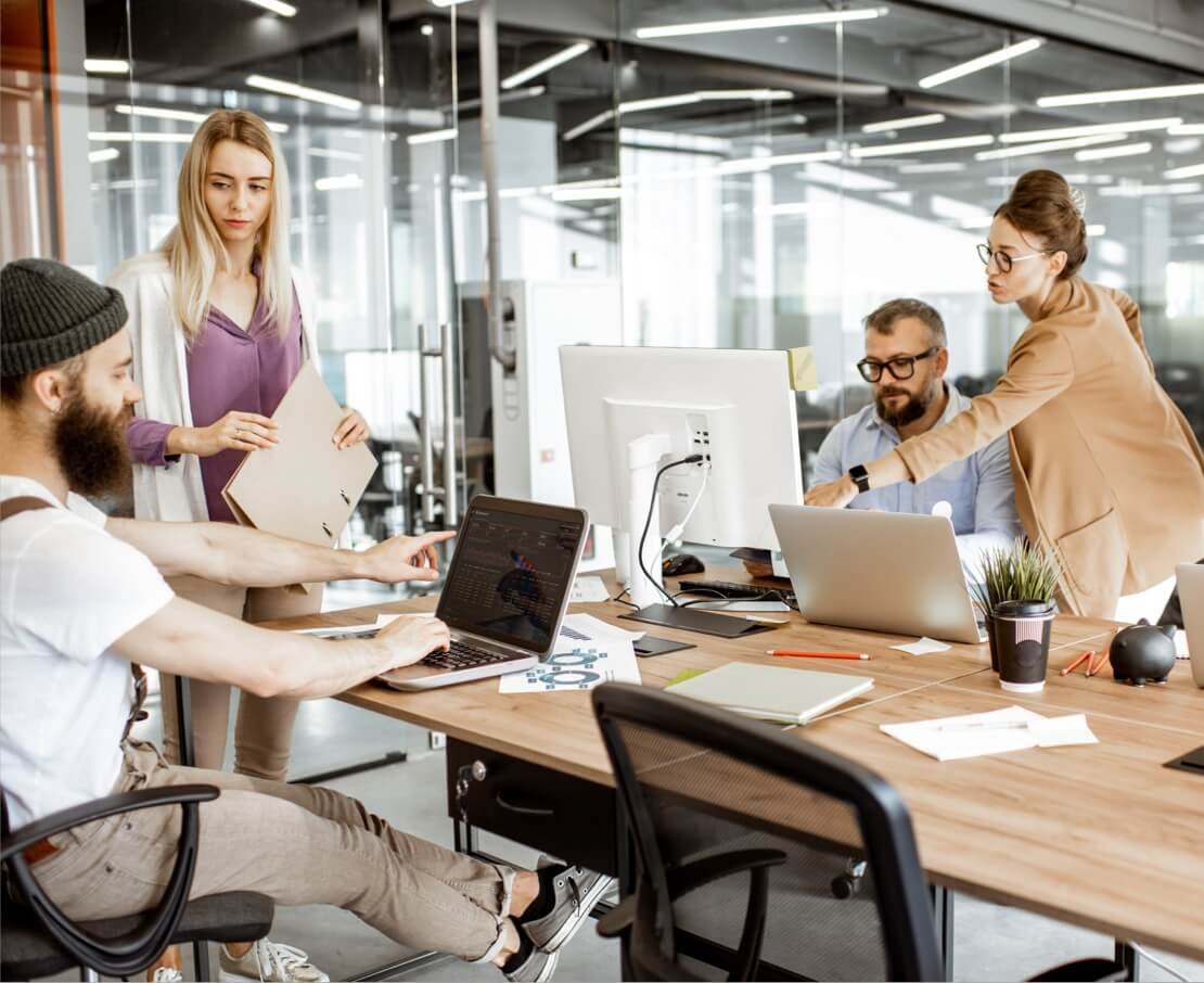 Team of four collaborating in a modern open-plan office around laptops and a desktop monitor.