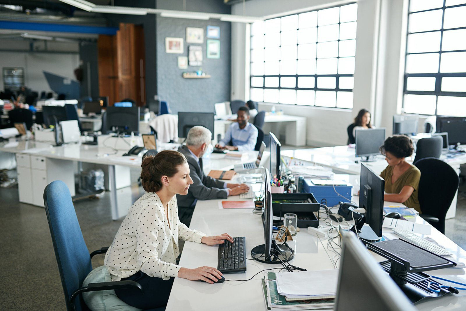 Open-plan office with multiple desks and coworkers working on computers near large grid windows.