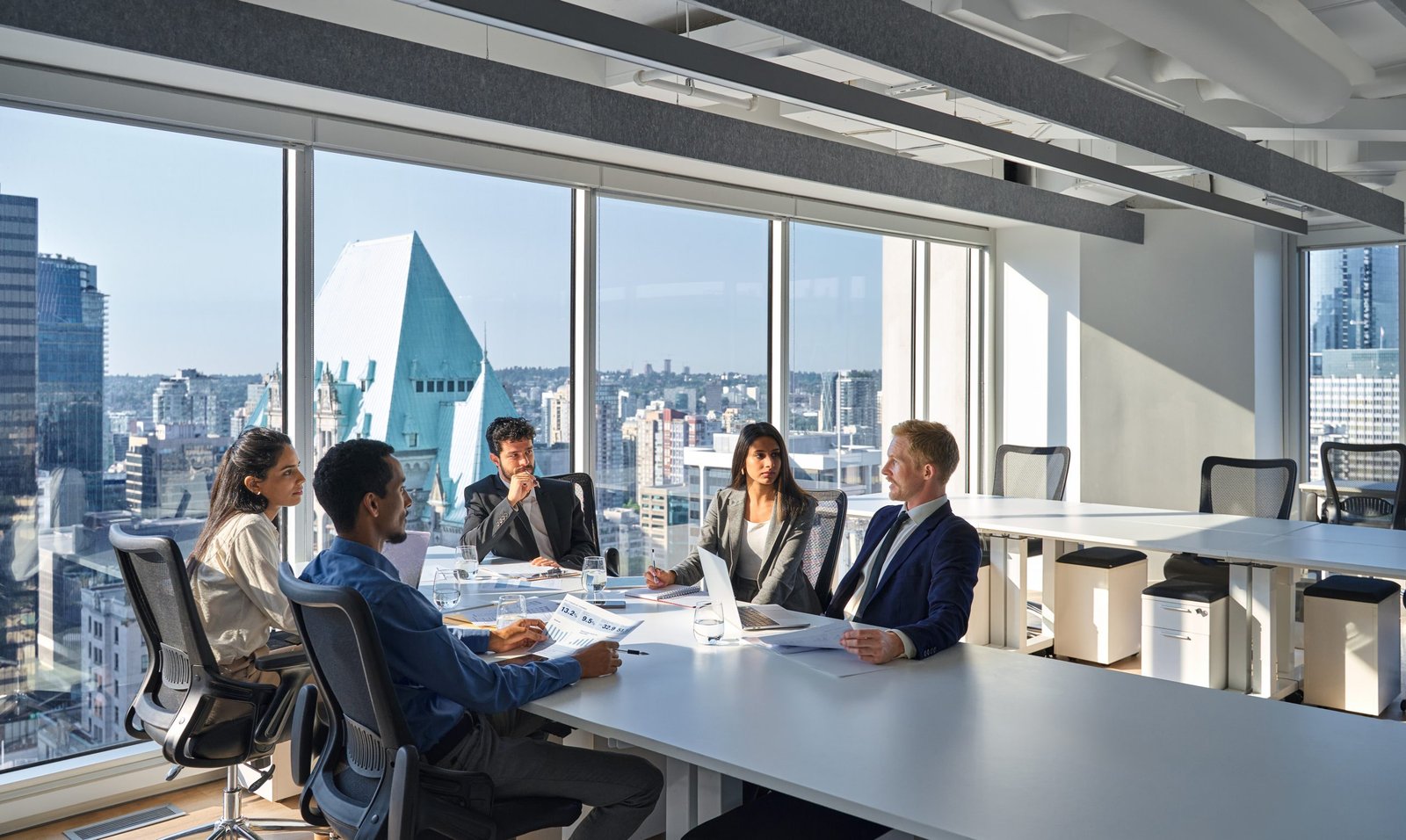 Business meeting in a glass-walled office with five professionals seated around a table, city skyline visible through large windows.