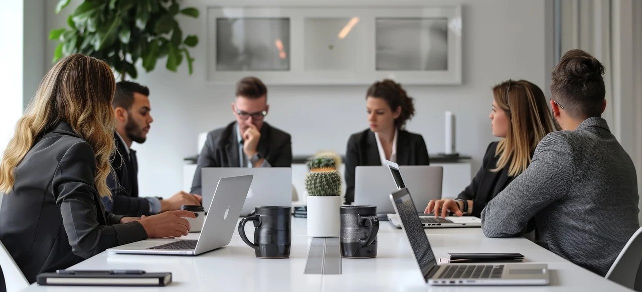 Six professionals sit around a white conference table in a bright office, each with a laptop, discussing together with coffee mugs nearby.