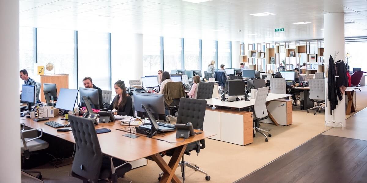 Open-plan office with rows of desks, computers, and employees working at their stations in a bright, modern space.