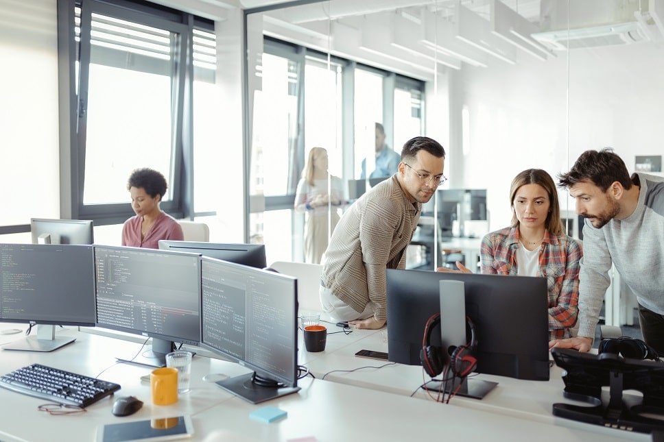 Team of four colleagues gathered around multiple computer monitors on a bright open-plan office, collaborating.