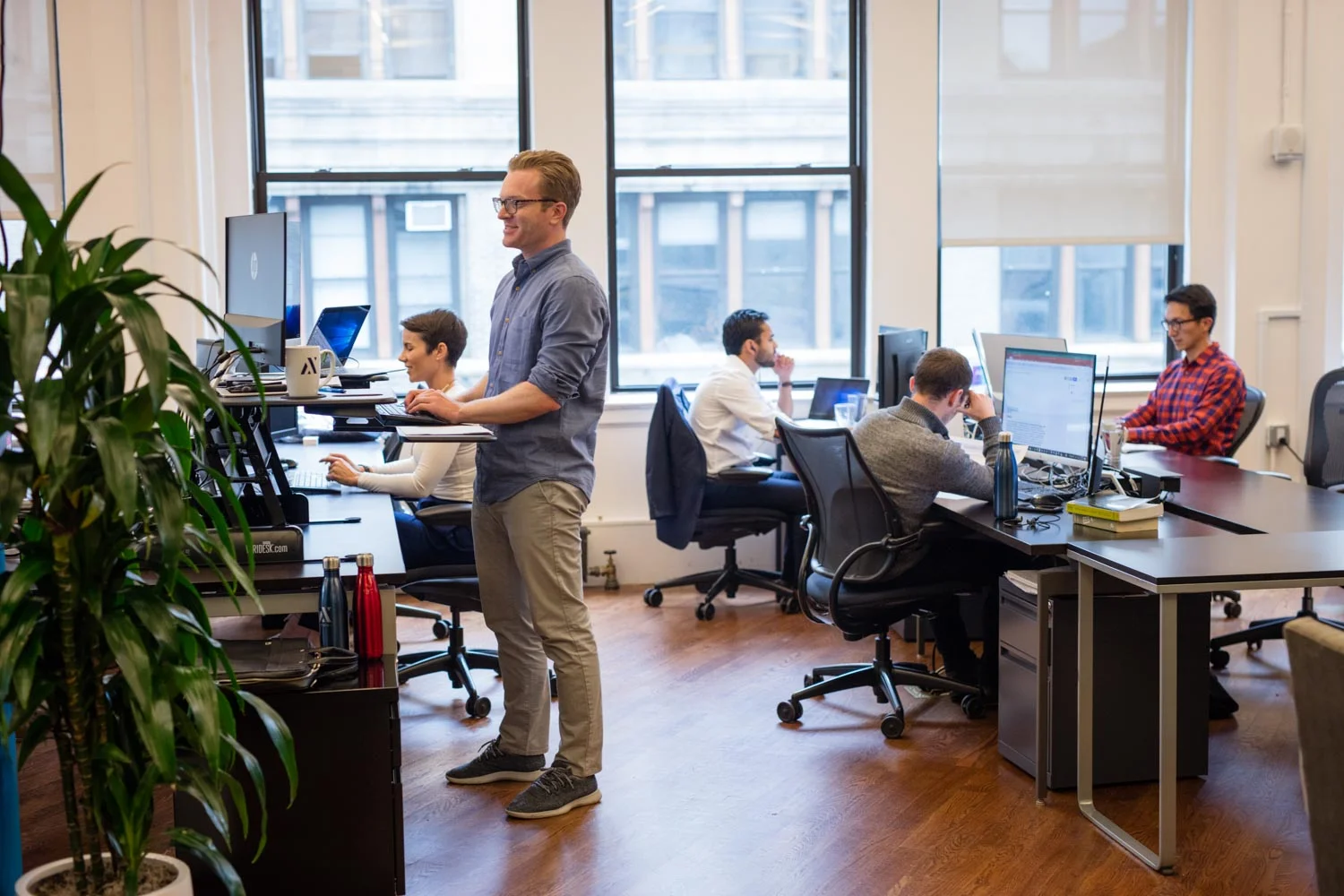 Office coworking space with several employees at desks and a man standing at a workstation with a laptop tray, smiling as he works.