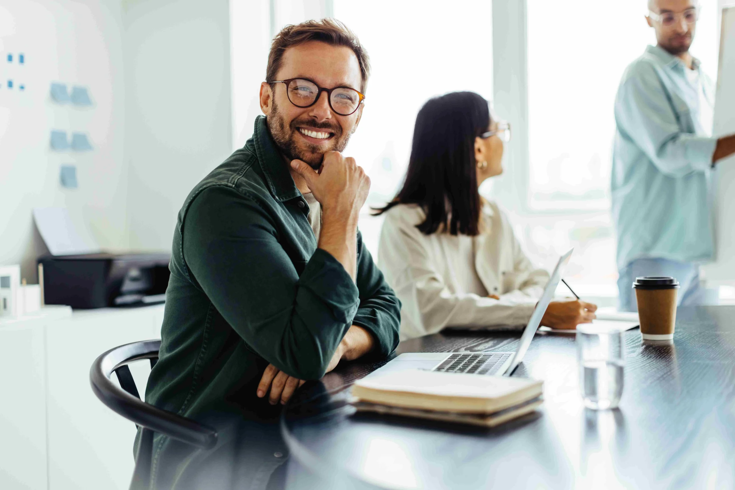 Smiling man with glasses sits at a desk in a bright office, coworkers in the background working.