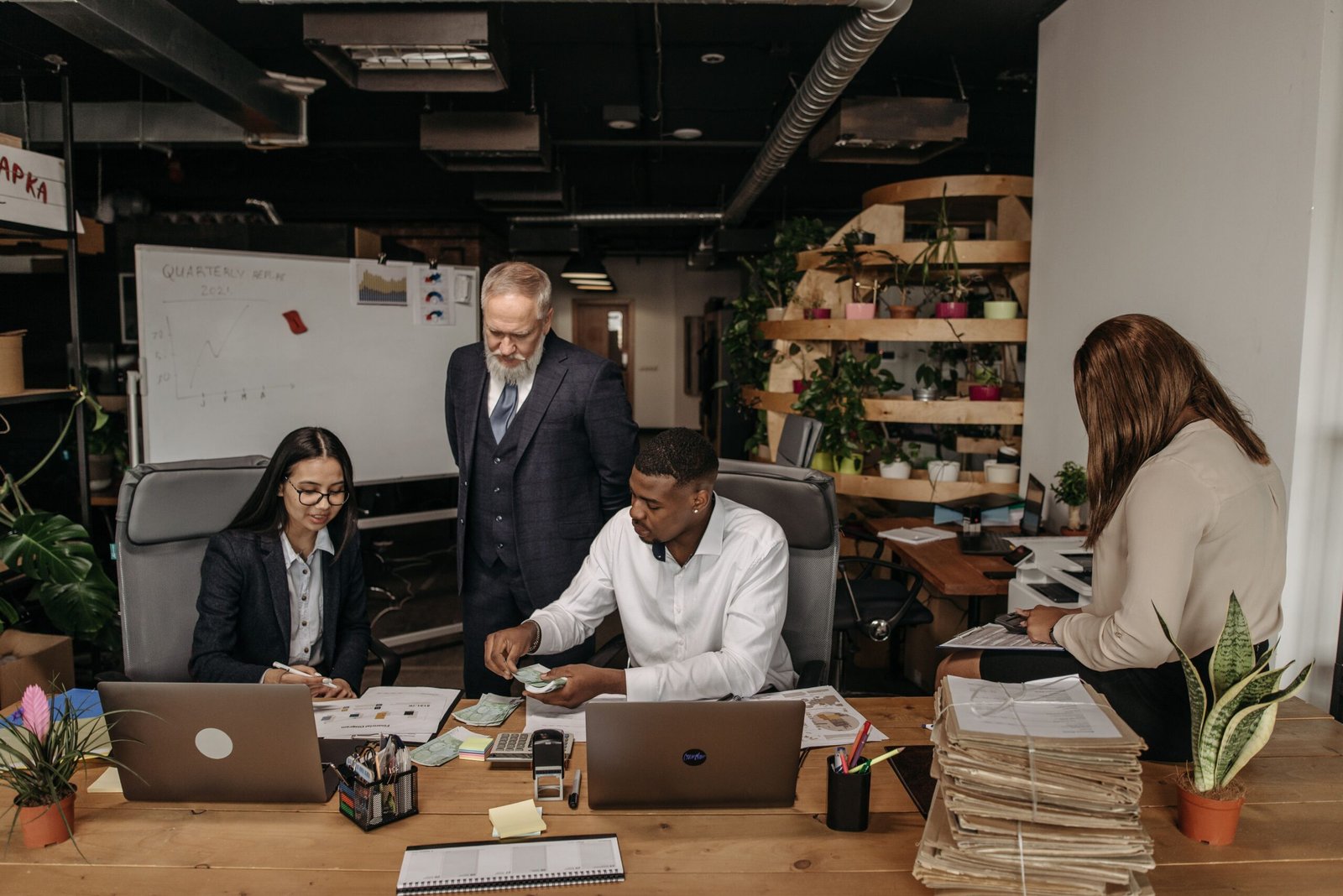 Team of professionals around a wooden table reviewing documents and laptops in a modern office. A whiteboard and greenery are in the background.
