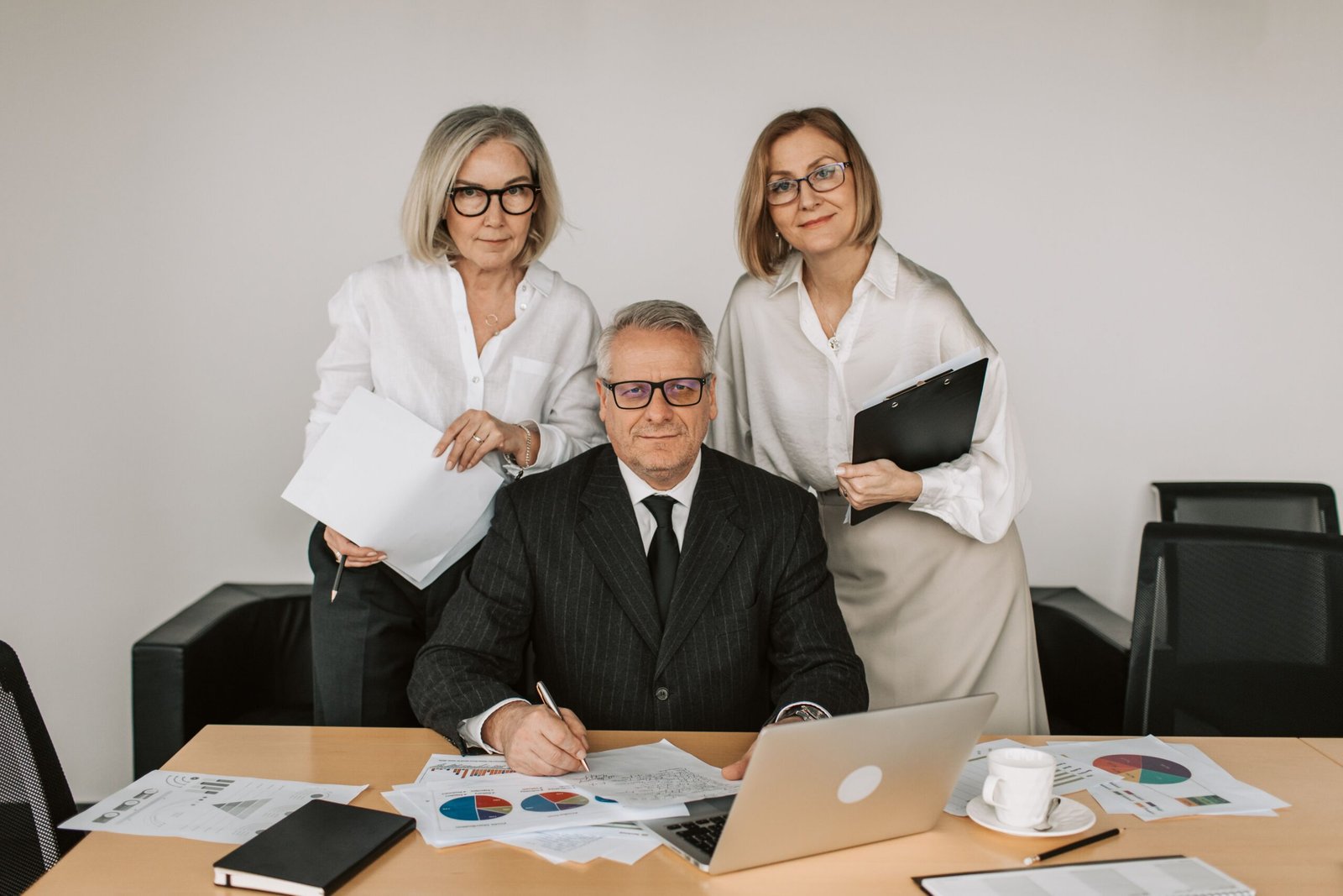 Three professionals posing in a modern office: a man in a suit sits at a desk signing papers while two women stand behind him with folders and documents.