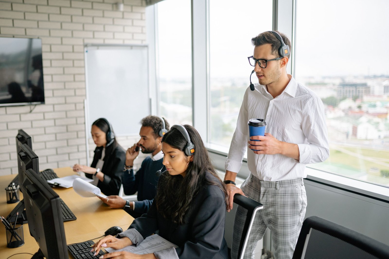 Team of customer service agents at computers wearing headsets; a supervisor stands with a blue coffee cup nearby in a bright office.